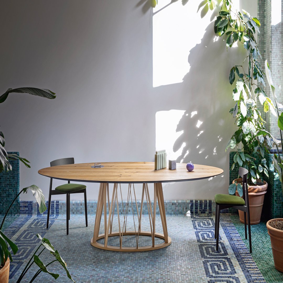 Round wooden dining table with natural wood finish, set on a geometric patterned blue and green tiled floor.