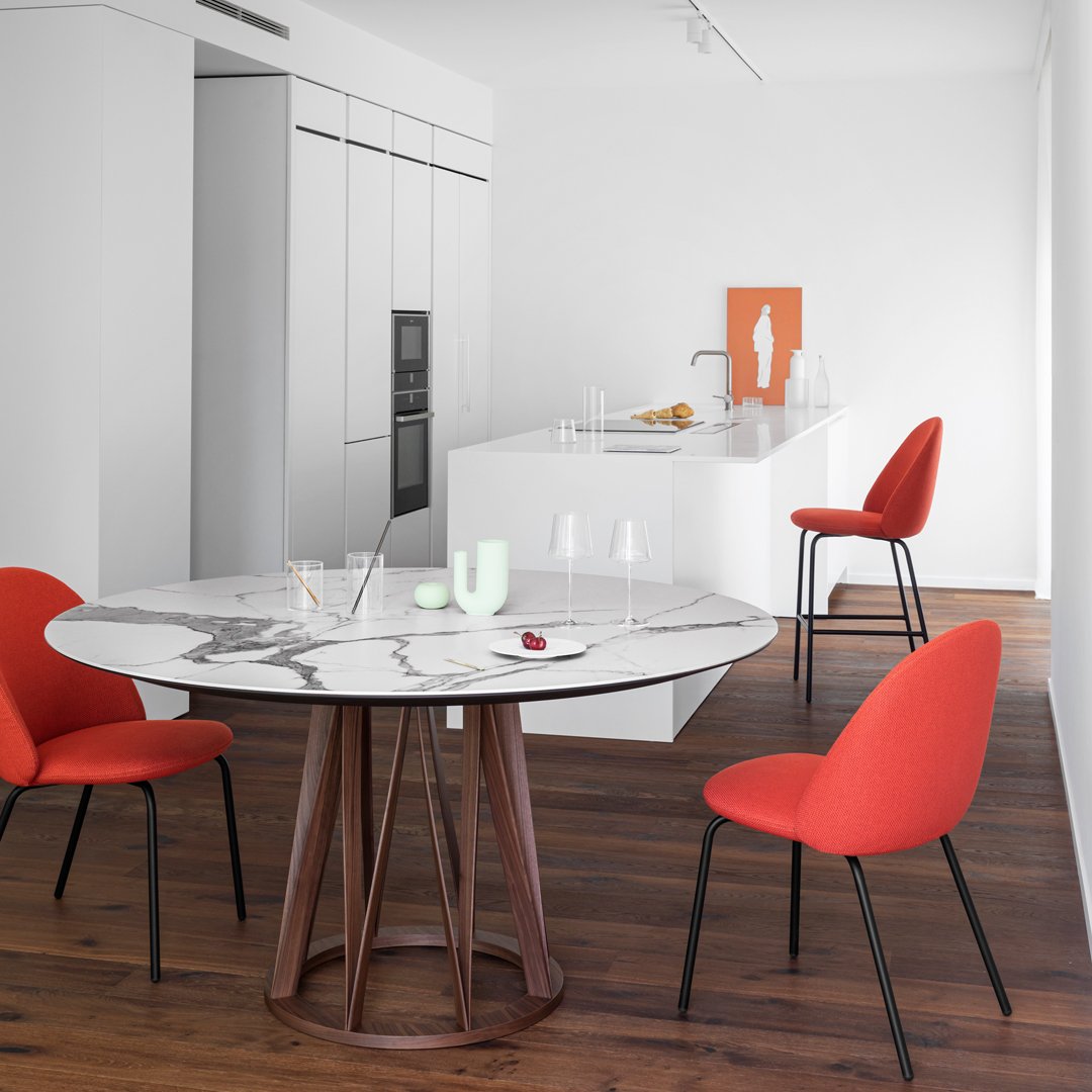 Round marble dining table with wood base, complemented by vivid red upholstered chairs against a modern white kitchen backdrop.
