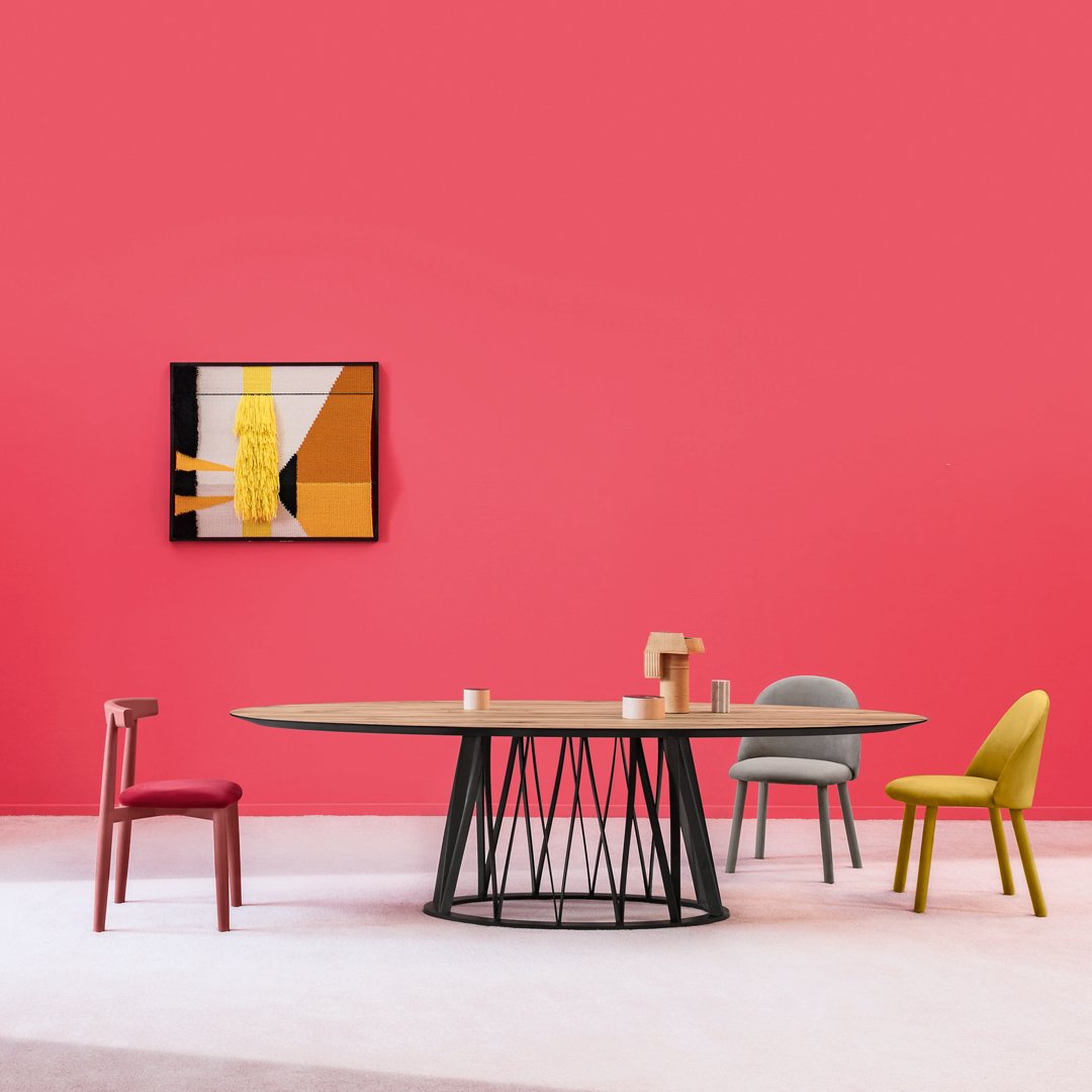 Modern dining setup featuring a round wood table, colorful chairs in gray, yellow, green, and red against a vibrant pink wall.