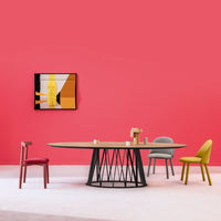 Modern dining setup featuring a round wood table, colorful chairs in gray, yellow, green, and red against a vibrant pink wall.