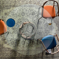 Transparent round table with patterned gray rug, featuring orange and blue chairs for a modern, stylish look.