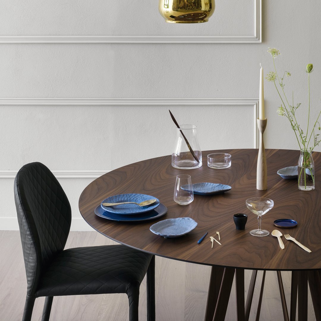 Elegant round wood table set with blue patterned plates, black textured chair, and minimalist glassware.