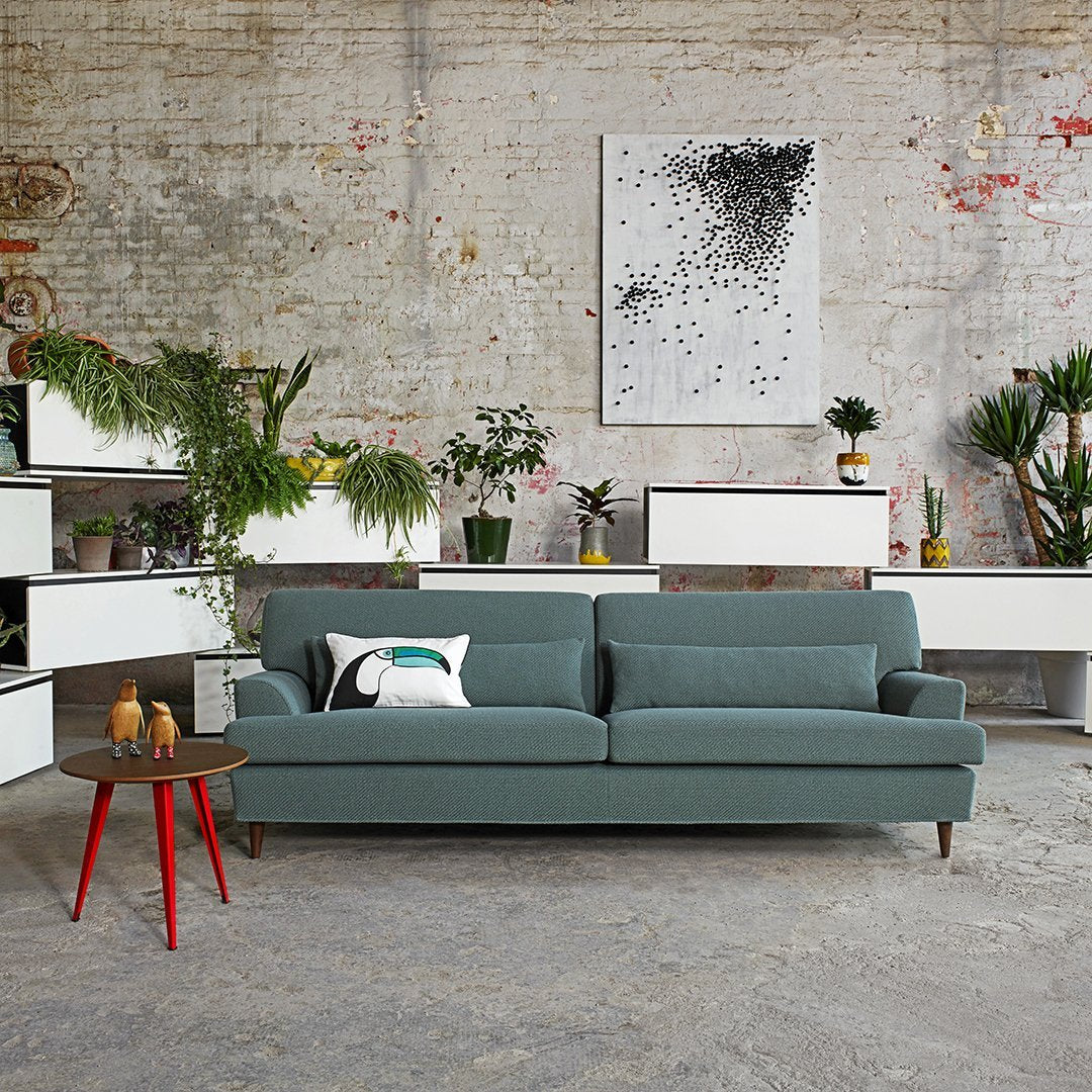 Teal sofa with a minimalist design, accented by red-legged side table and greenery against a textured gray wall.