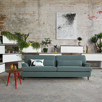 Teal sofa with a minimalist design, accented by red-legged side table and greenery against a textured gray wall.