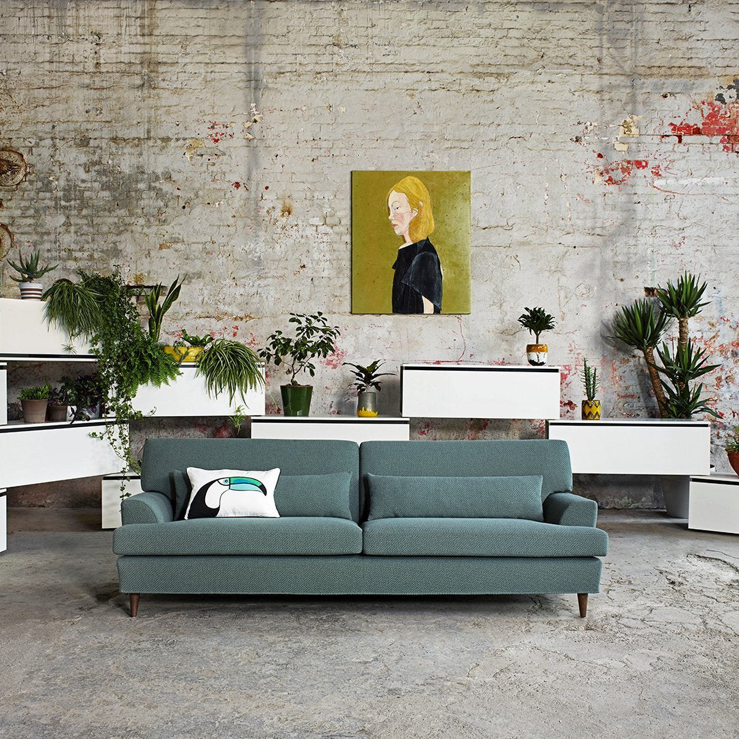 Teal modern sofa with minimalistic design, complemented by green plants and neutral-toned decor in an industrial setting.