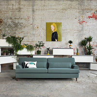 Teal modern sofa with minimalistic design, complemented by green plants and neutral-toned decor in an industrial setting.