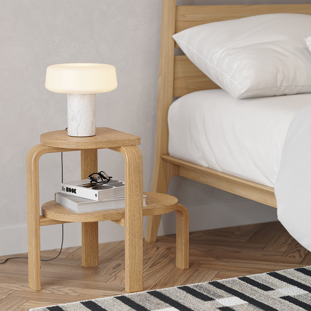 Natural wood nightstand with light-colored lamp, paired with a cozy white bed and a striped black-and-white rug.