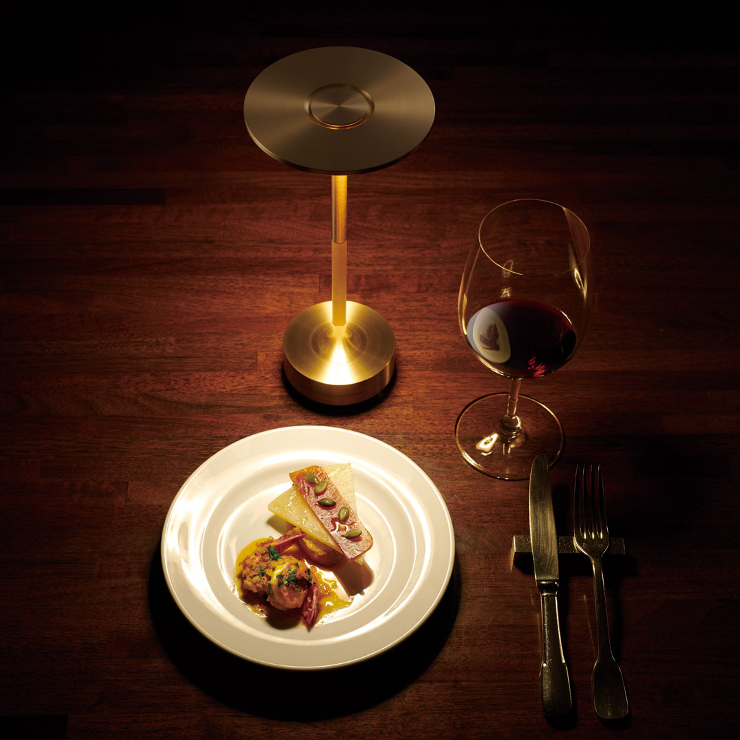 Elegant gold table lamp with a warm glow; contrasting white plate and rich red wine against dark wood background.