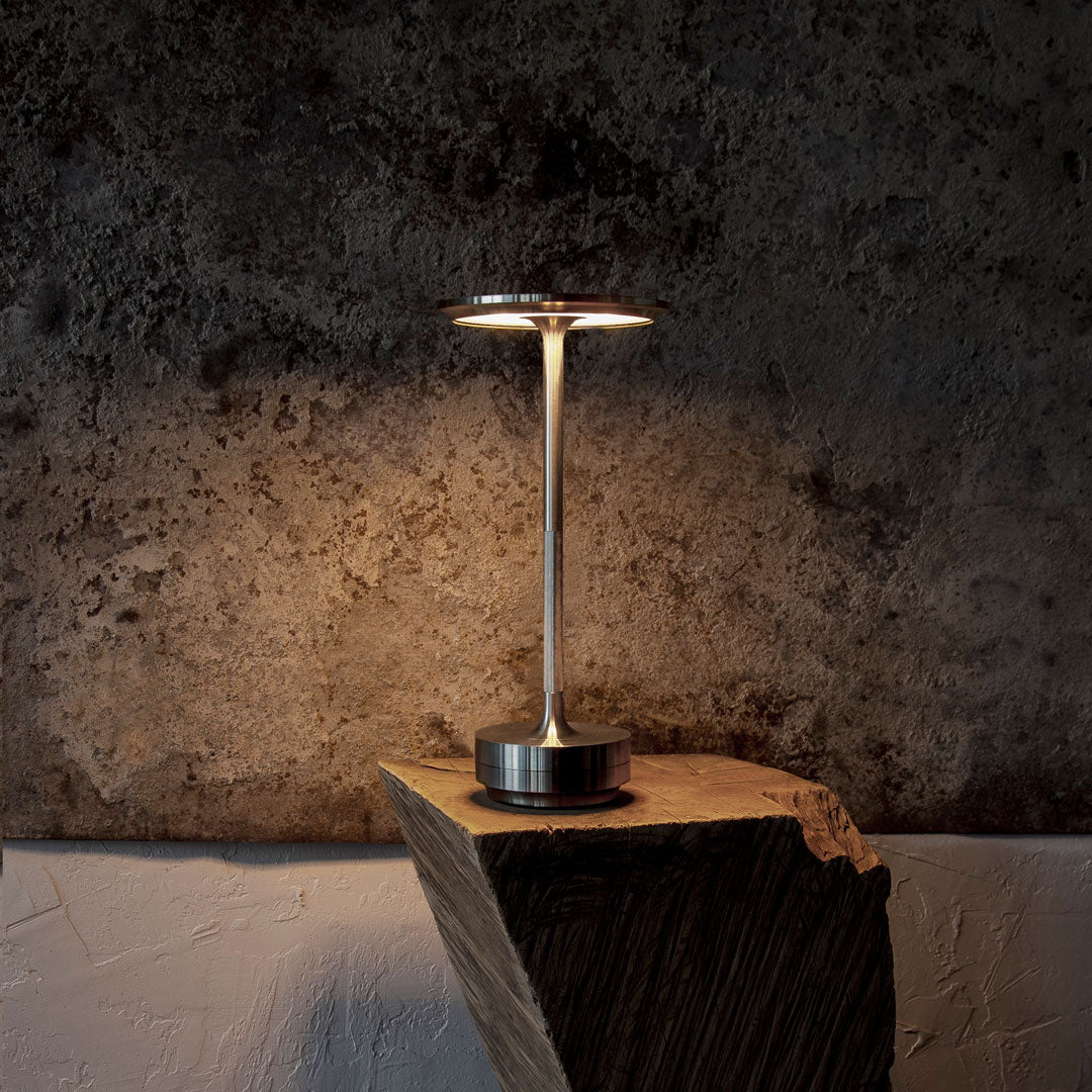Sleek silver lamp with a modern design, set against a textured dark stone wall and light wooden surface.