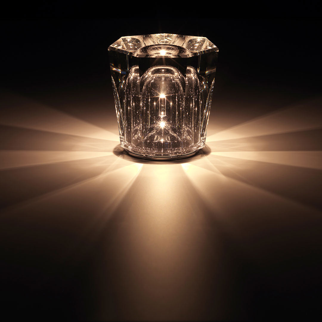 Crystal glass candle holder with intricate patterns, casting warm golden light and shadows against a dark backdrop.