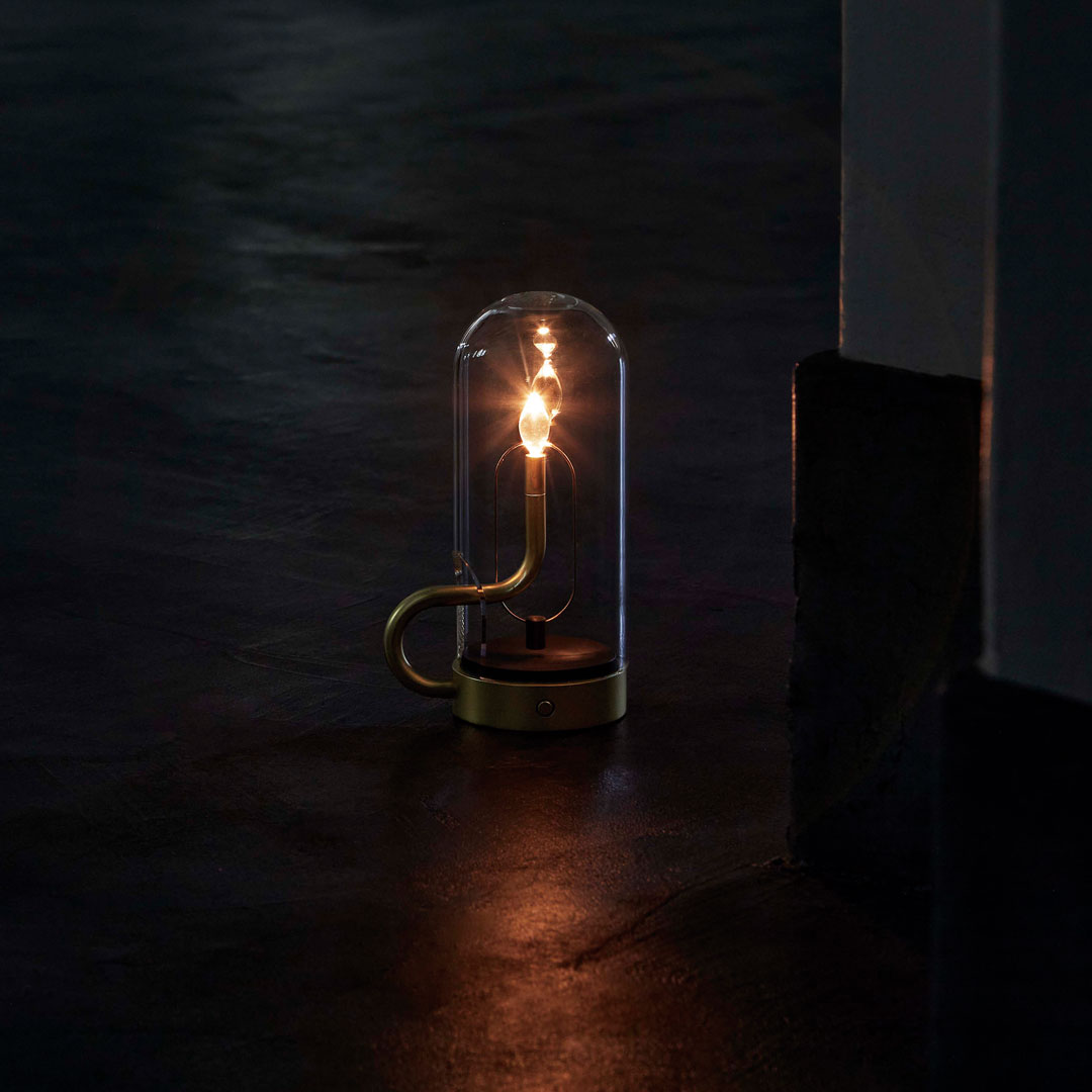 Clear glass lamp with a warm white bulb, featuring a sleek brass base and minimalist design on a dark surface.