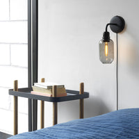 "Black wall sconce with a glass bulb beside a navy blue quilt and a two-tone black and wood side table."