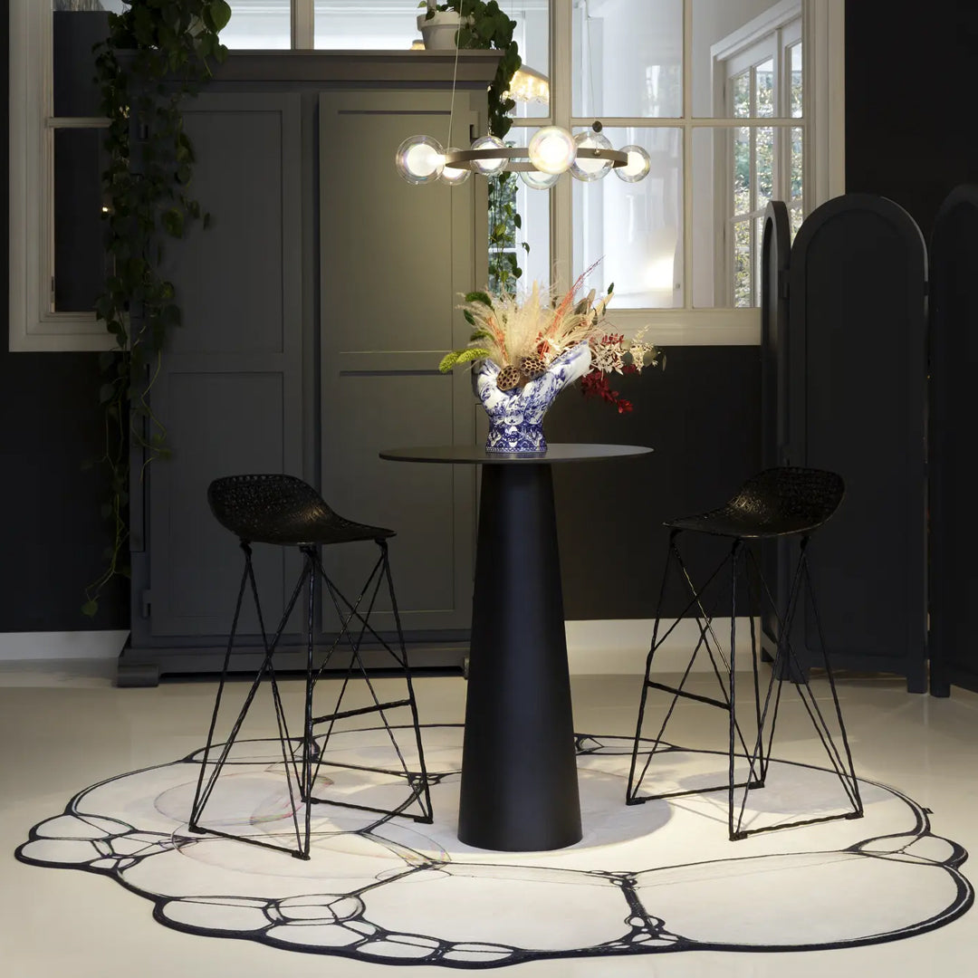 Black round table, clear vase with colorful flowers, black bar stools, and a white rug with a black abstract pattern.