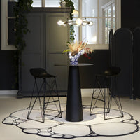 Black round table, clear vase with colorful flowers, black bar stools, and a white rug with a black abstract pattern.