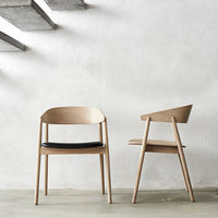 Two minimalist wooden chairs: light natural wood with black cushions, set against a textured white wall.