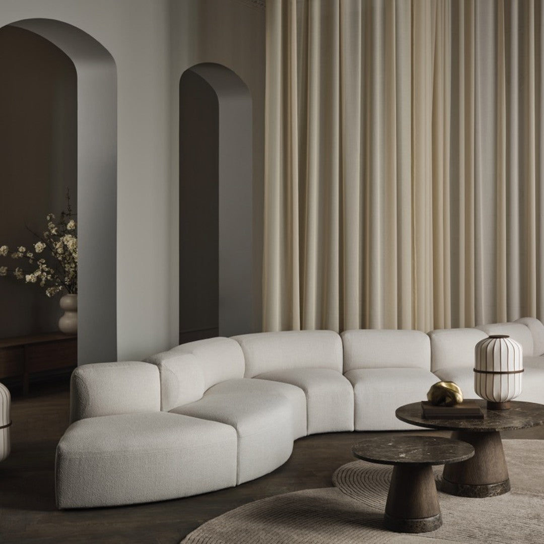 Curved beige sofa with textured upholstery, paired with dark wooden tables and light cream curtains. Modern neutral palette.