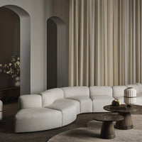 Curved beige sofa with textured upholstery, paired with dark wooden tables and light cream curtains. Modern neutral palette.