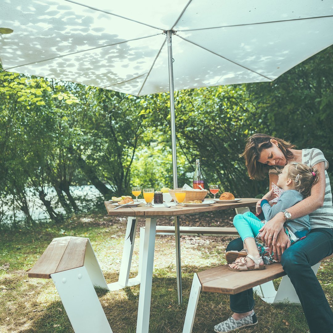 "Bright white umbrella with green trees; wooden table and benches; soft pastel colors create a serene outdoor setting."