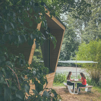 "Wooden cabin with angular design, surrounded by green foliage, featuring a white picnic table under a light green umbrella."