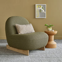 Olive green chair with textured fabric, beige pillow, and natural wood accents against a muted mustard wall and gray rug.