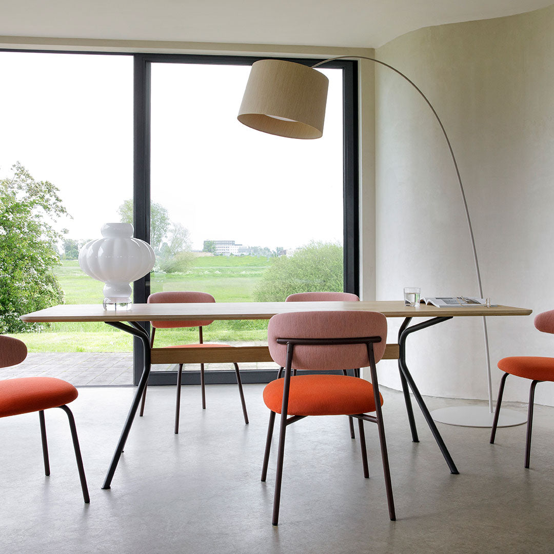 Modern dining room with orange and pink chairs, light wooden table, and a beige lamp against a minimalist backdrop.