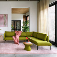Lush green sectional sofa with light pink throw and geometric side table on soft pink rug in modern living space.
