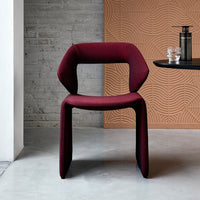 Burgundy upholstered chair with smooth curves, set against a textured beige wall. Minimalist modern design.