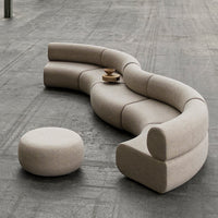 Curved beige sofa with smooth lines and a matching round pouf on a textured gray floor. Elegant minimalist design.