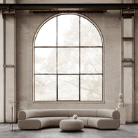 Curved beige sofa and round coffee table against a minimalist, light-filled backdrop with large arched window.