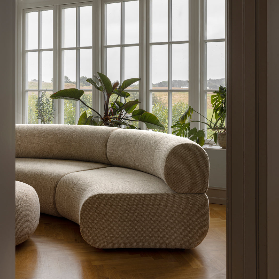 Beige curved sofa with a smooth texture, set against large windows and greenery. Natural light enhances its minimalist design.
