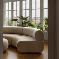 Beige curved sofa with a smooth texture, set against large windows and greenery. Natural light enhances its minimalist design.