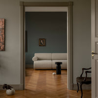 Modern beige sofa in a green wall setting with herringbone wooden flooring and contrasting dark accent pieces.