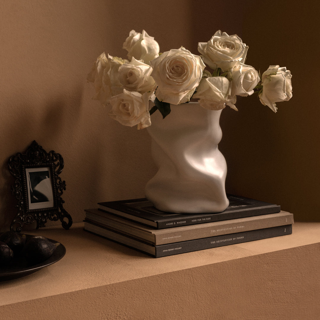 White textured vase holding cream roses atop stacked black and beige books against a warm, earthy background.