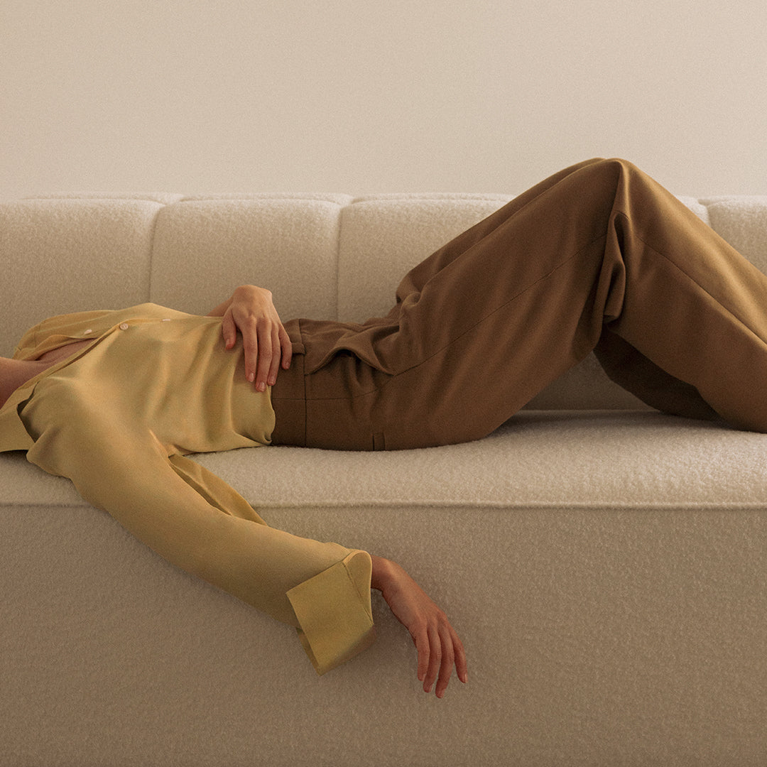 Light yellow blouse paired with brown wide-leg pants, set against a neutral-toned sofa. Elegant and modern colors.