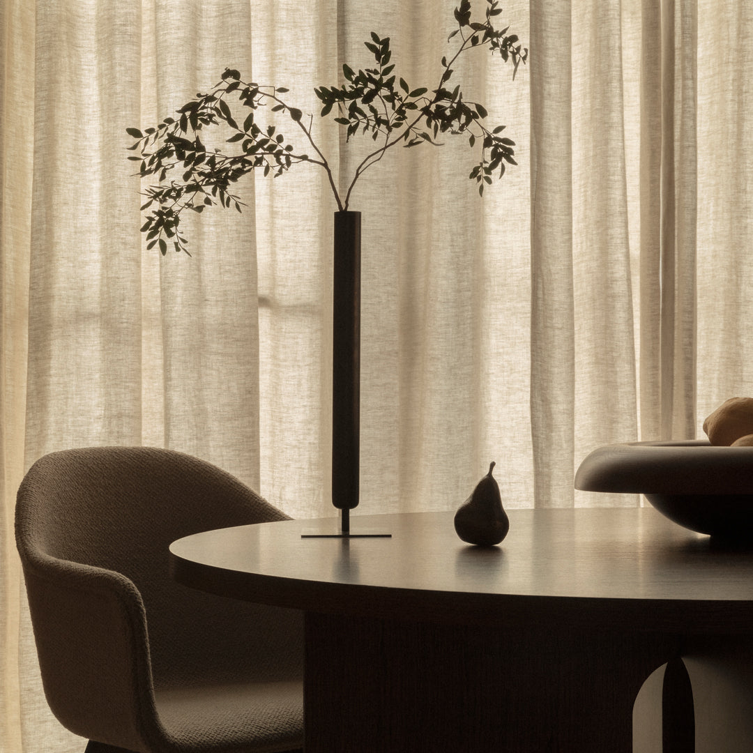 Black vase with greenery on a round dark wood table, complemented by soft beige curtains and a subtle light palette.