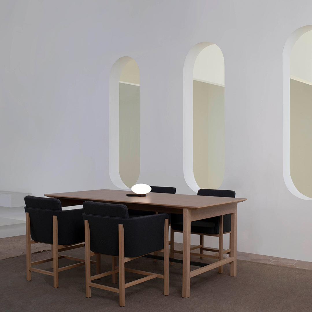 Modern wooden dining table with black upholstered chairs, set against a neutral wall and soft, curved window patterns.