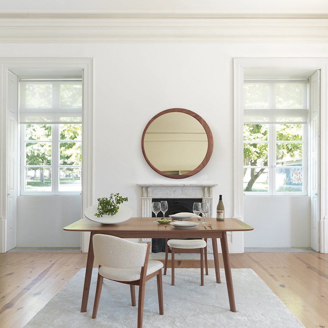 Modern dining room featuring a round wooden mirror, neutral beige chairs, and a light wood table on a soft gray rug.