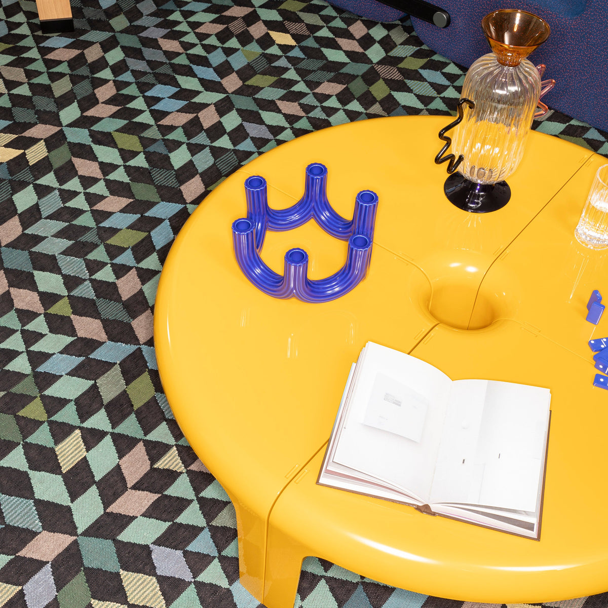 Bright yellow round table with blue plastic decor on a geometric patterned rug featuring teal, brown, and black hues.