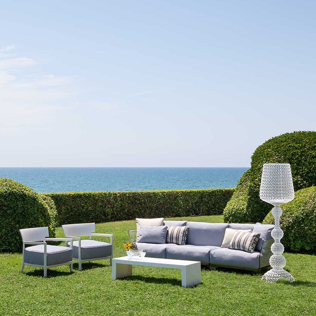 White outdoor sofa set with striped cushions, accompanied by a decorative lamp in an open green space by the sea.