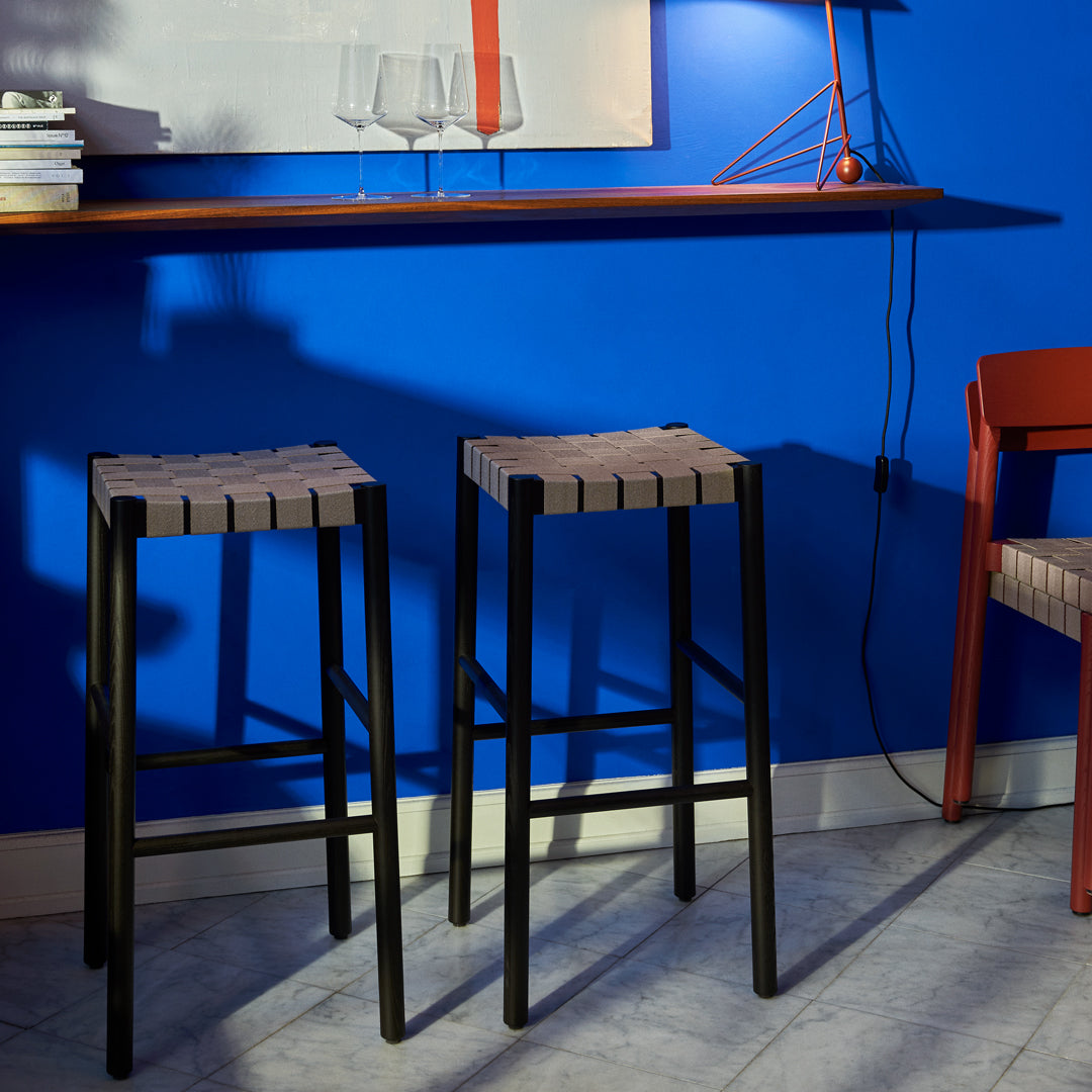 "Two black-framed bar stools with wooden slatted seats against a blue wall, complemented by a red chair."