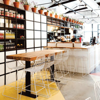 Minimalist café with a wooden table, white wire chairs, and vibrant potted plants against a clean, neutral backdrop.