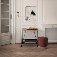 Black lamp, gray metal cart, textured red and beige pouf, with a neutral wall and herringbone wood floor pattern.