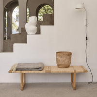 Natural wood bench with woven pattern, grey throws, and a textured wicker basket against a minimalist backdrop.