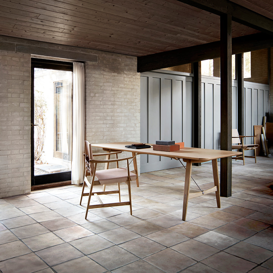 Modern wooden table with light wood finish, paired with soft pink chair, set against textured gray walls and tiled floor.