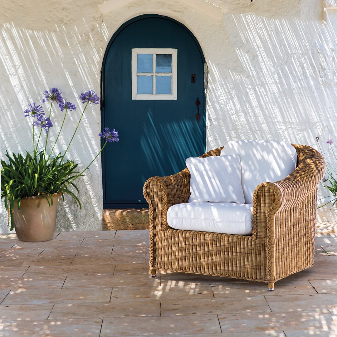 Natural woven rattan chair with white cushions against a textured beige wall and blue door. Soft floral accents nearby.