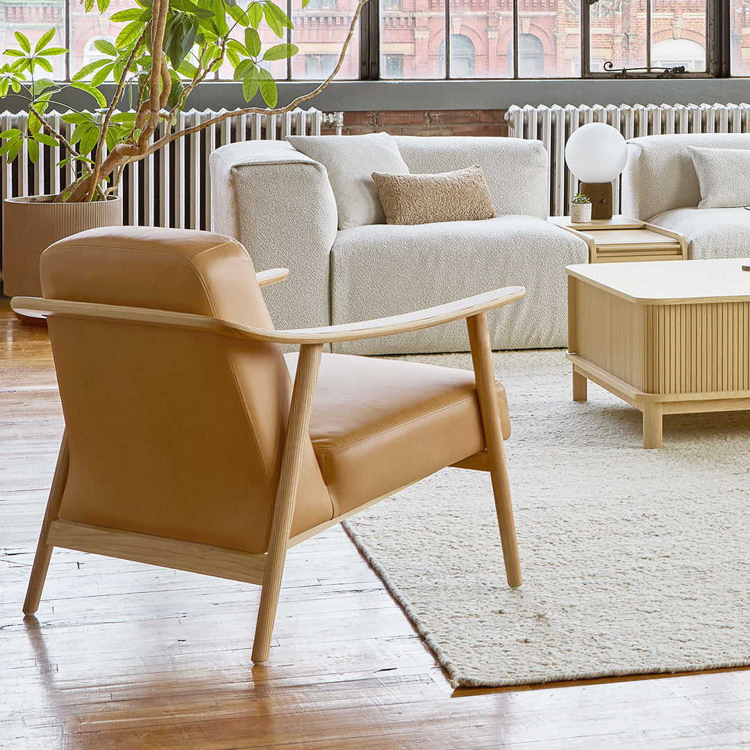 Tan leather armchair with light wood accents, set against neutral textured upholstery and a soft, cream rug.