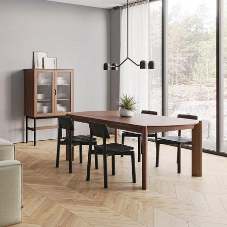 Modern wooden dining table with black chairs, set against a light gray wall and herringbone-patterned wood floor.
