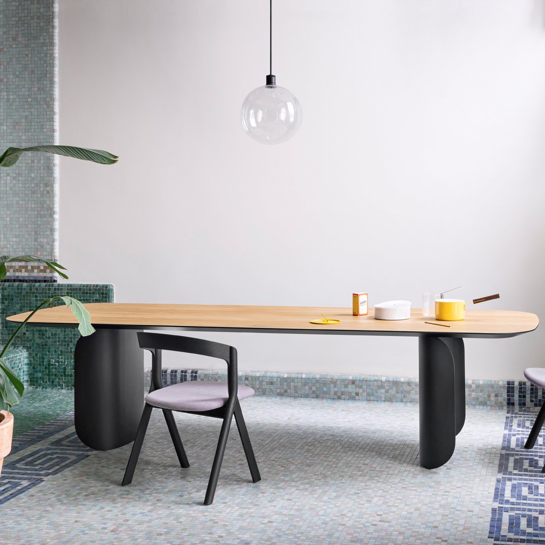 Light wood tabletop with smooth black legs, against a patterned blue floor and minimalistic decor.