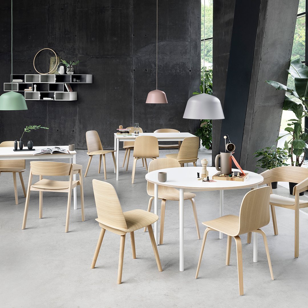 Modern dining area featuring light wood chairs, white tables, and muted green and grey lamps against a dark wall.
