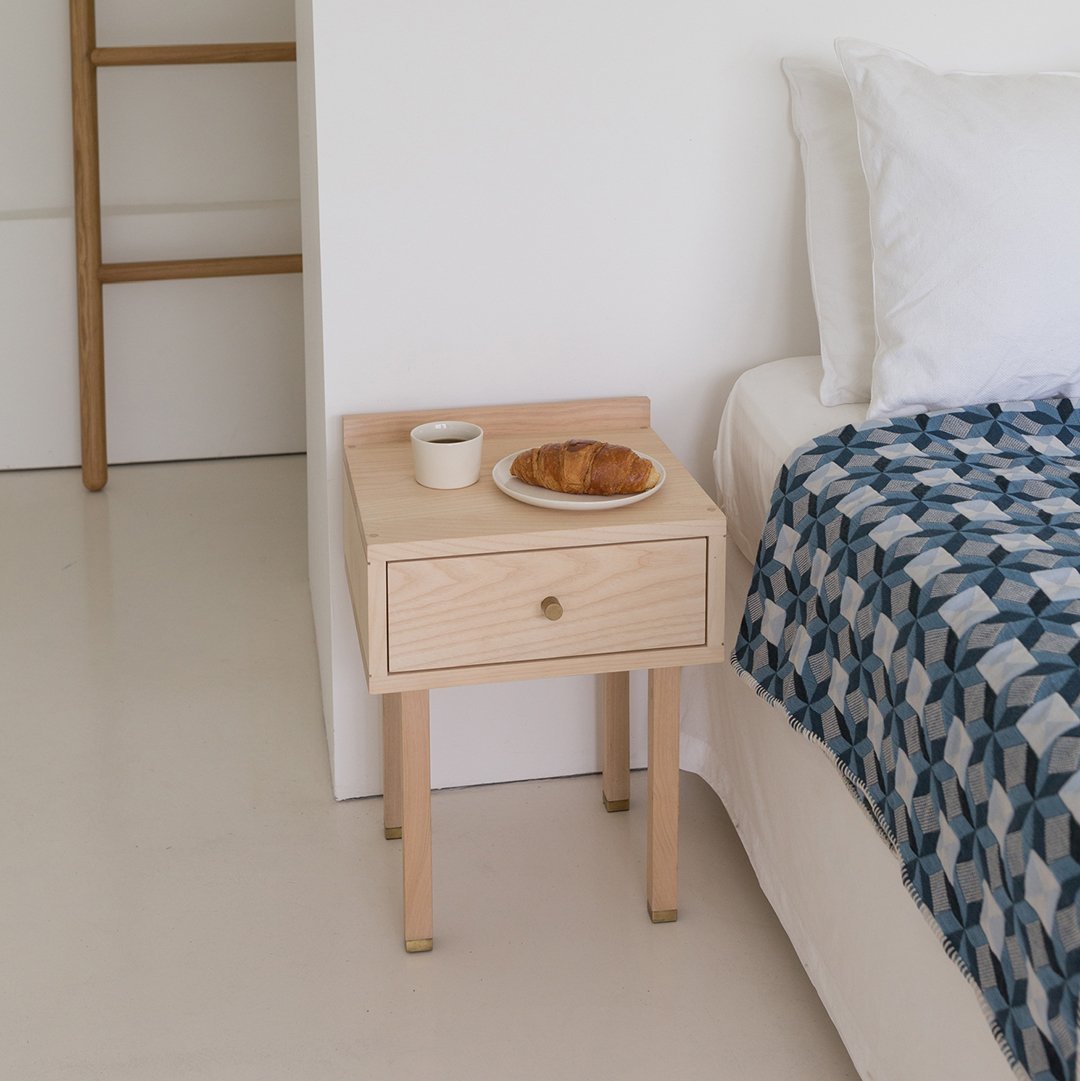 Light wood nightstand with a single drawer, complemented by a blue patterned quilt on the bed.